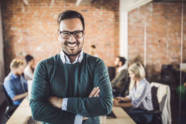 “Smiling man with glasses standing confidently in modern office with people working behind him.”