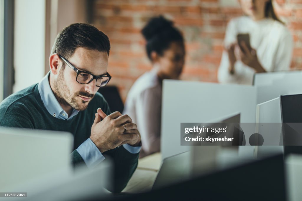 “Focused man with glasses working at computer in modern office, colleagues collaborating behind him.”