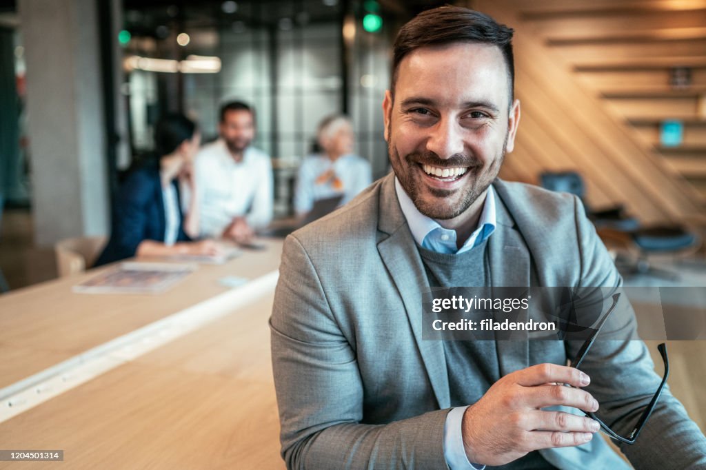 “Smiling businessman holding glasses, sitting at desk in modern office, colleagues talking in background.”