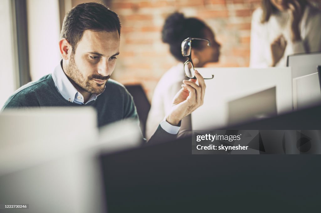 “Serious man holding glasses, concentrating at computer in modern office, colleagues working in background.”