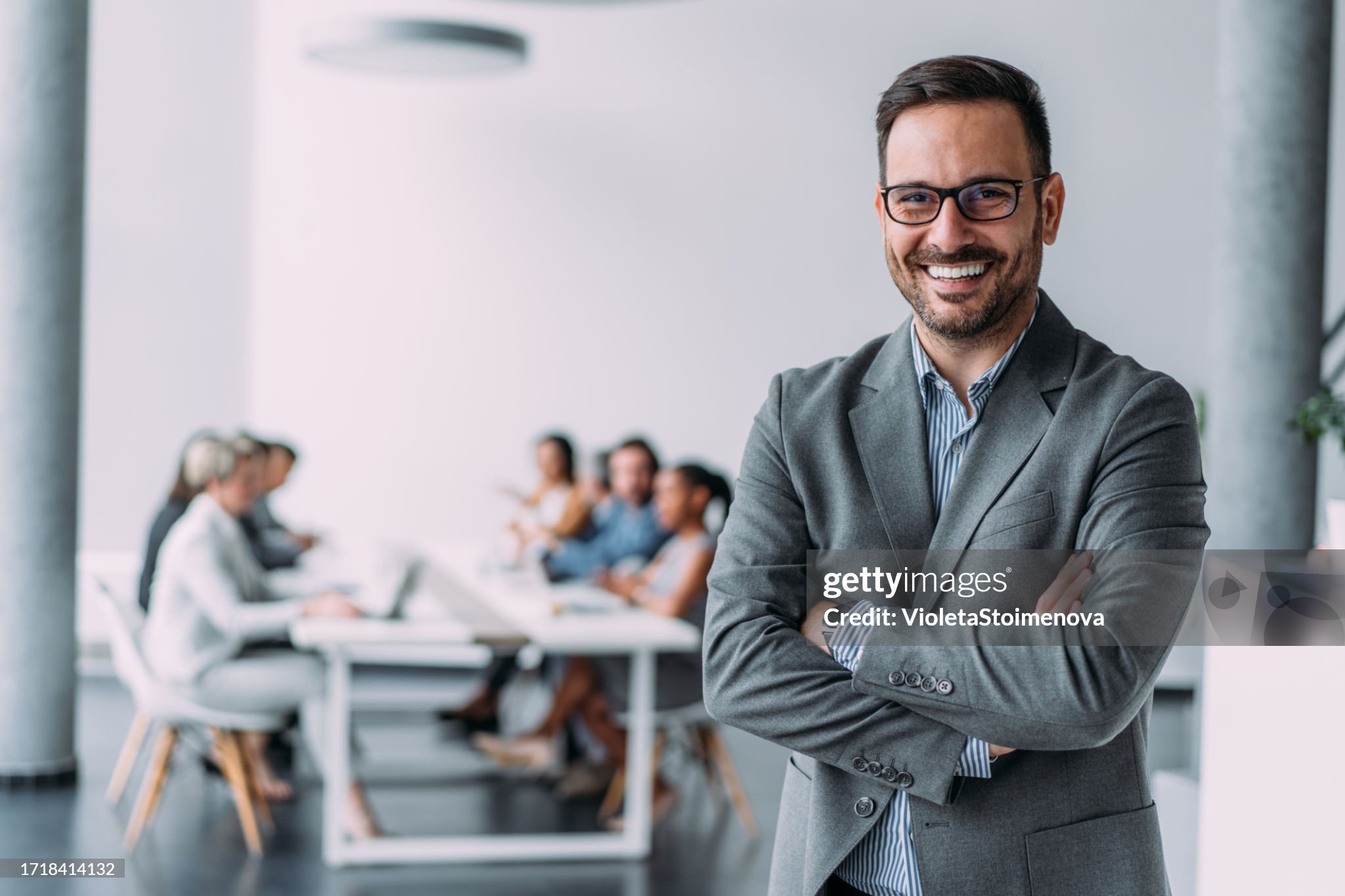 “Smiling man in suit standing in front of a meeting room with people at a table.”